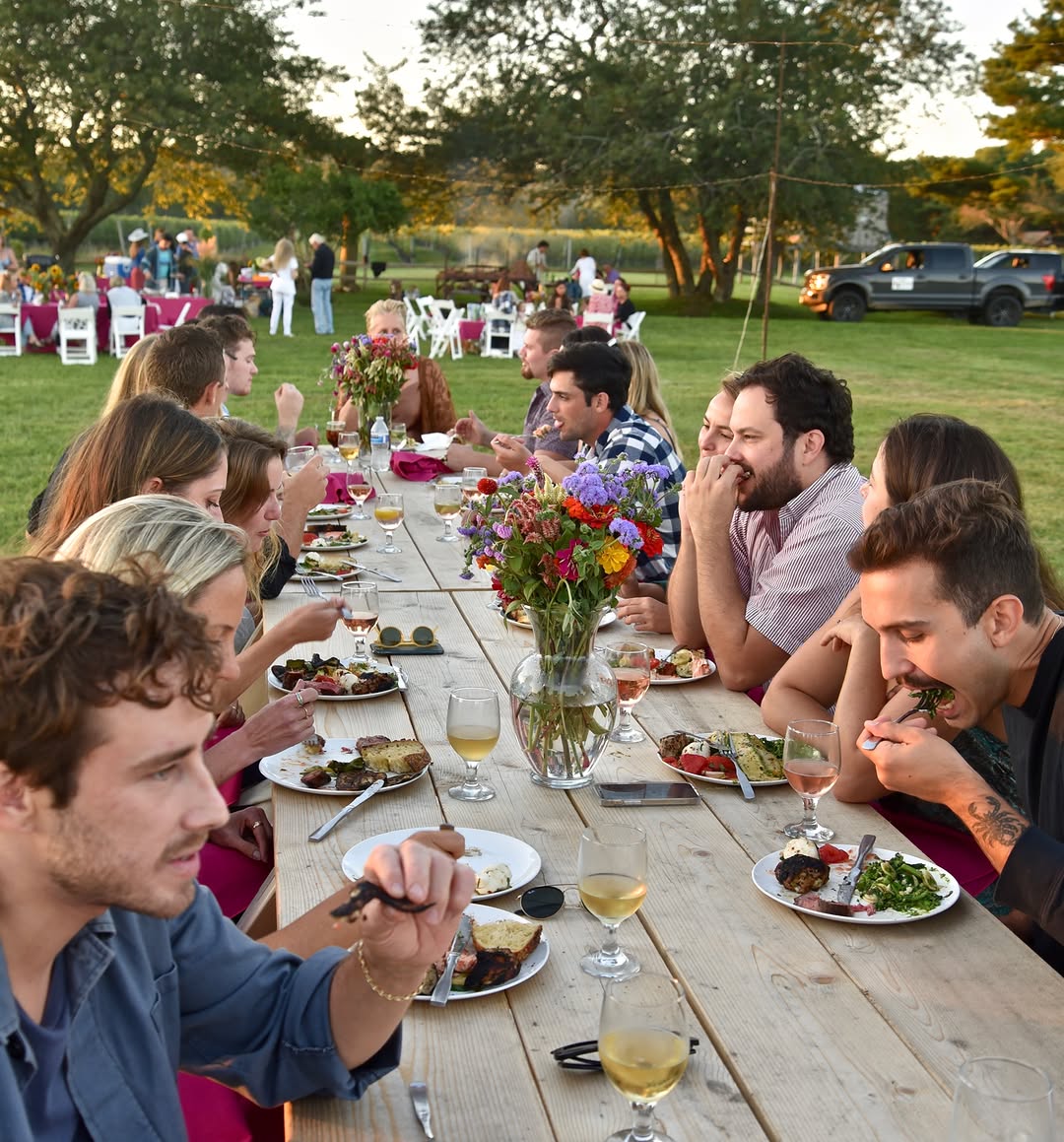 A long farm table dinner on the lawn at golden hour, flowers and wine glasses down the middle